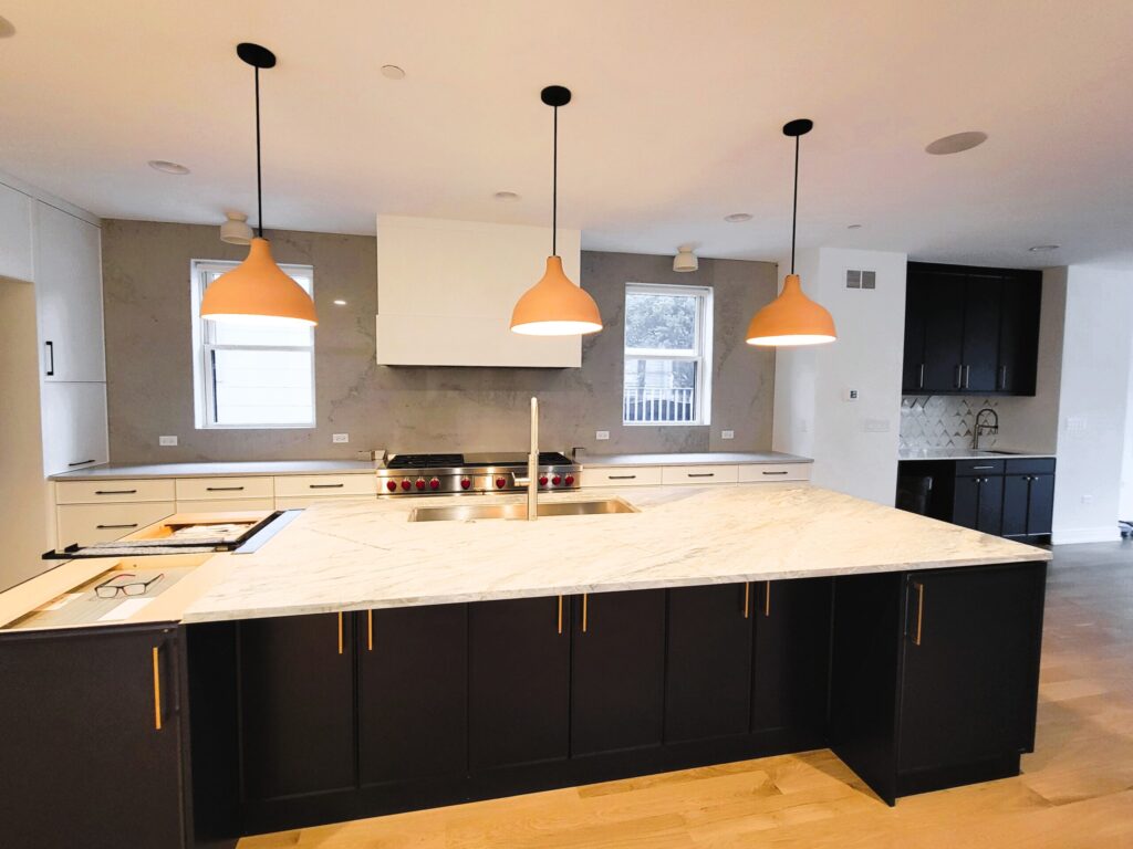 A contemporary kitchen centered around a massive island with a white veined stone countertop, built-in sink, and dark black cabinetry with gold drawer pulls. Three large, matte terracotta-colored bell pendant lights hang above the workspace. The rear wall features white base cabinets, a professional-grade stainless steel range with red knobs, and a full-height grey stone backsplash between two small windows. To the right, a separate wet bar area with black cabinets and a geometric backsplash is visible.