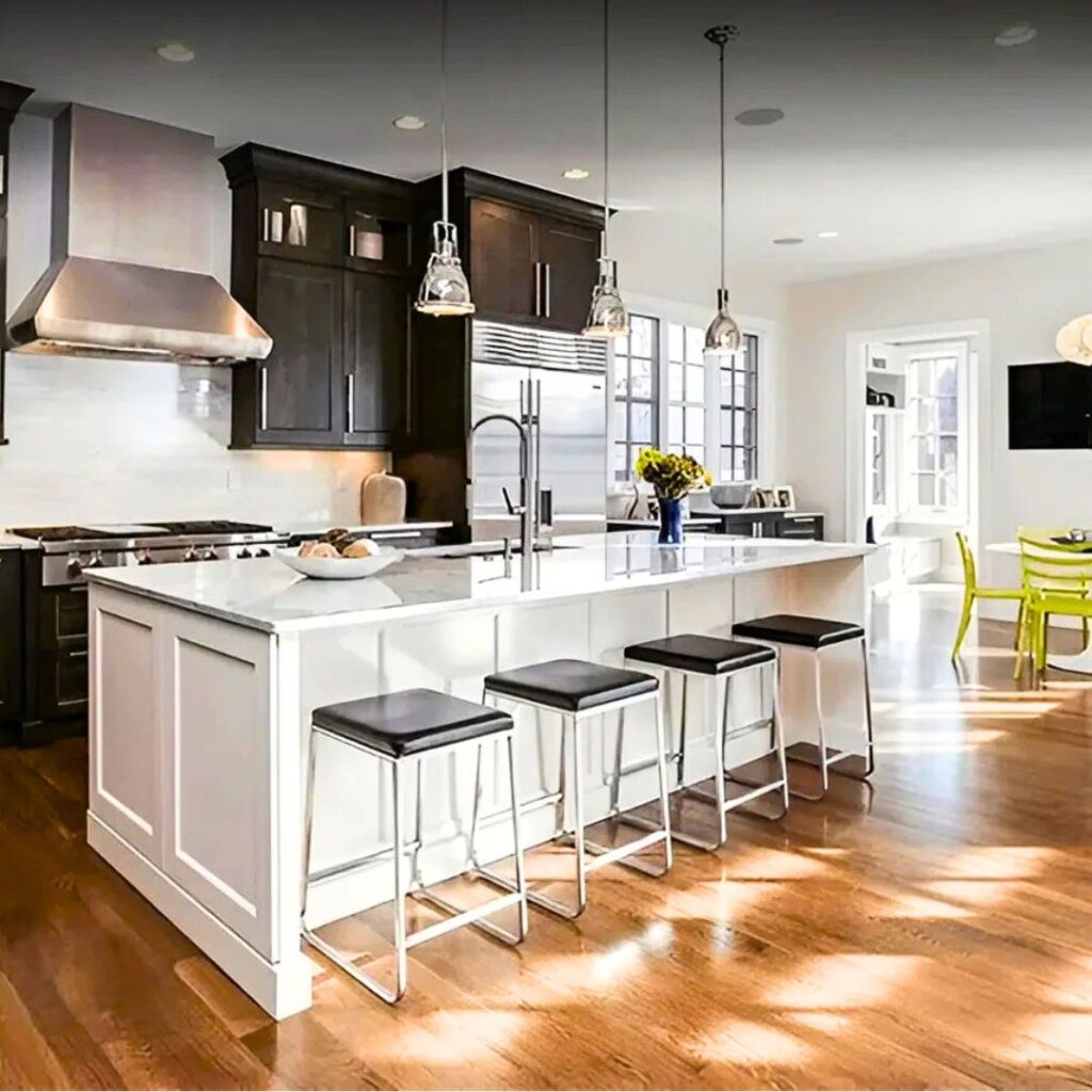 A spacious, modern kitchen featuring a large white island with a white marble countertop and four backless bar stools with chrome legs and black seats. Contrasting dark wood cabinetry lines the back wall, accompanied by stainless steel appliances, including a professional-grade range and large hood. Three glass pendant lights hang above the island, and sunlight streams across the medium-tone hardwood floors. To the right, an adjacent dining area is visible featuring bright lime-green chairs.